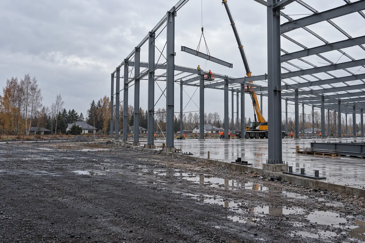 The steel frame of a large hall being assembled on a building site at the edge of a Polish town, with a crane lifting a beam